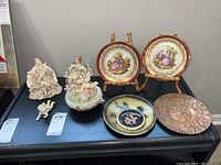 Photo of various hand-painted figurines, collector plates, covered dish, and decorative plates on a black surface