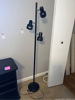Full view of black floor lamp standing next to wall and furniture, showing three adjustable light heads attached to a central pole with round base.