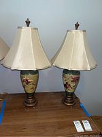 Two matching ceramic table lamps with beige hexagonal fabric shades, floral painted bodies, and ornate metal bases.