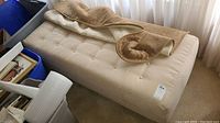 Photo showing cream-colored upholstered ottoman bench with button-tufted top and light brown throw blanket laid on top.