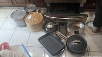 Overview of entire lot including pots, pans, steam basket, stainless steel kettle, metal mixing bowls, and bake pans arranged on floor in front of oven.