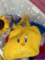 Photo showing an open plastic toy box containing various plush toys including a large yellow Tweety Bird plush and other colorful soft toys inside the box.