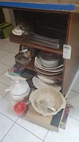 Full view of kitchenware lot including glass bakeware on shelving, tureens, teapot, cutting boards, and Pampered Chef bundt pan on floor