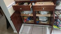 Open kitchen cabinet showing lower shelves and drawers containing Corningware dishes, utensils, and table linens.