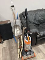 Photo of Bissell CleanView upright vacuum cleaner with orange and gray colors, a metal shovel with a D-handle, and a beige foldable small step stool next to a black couch.