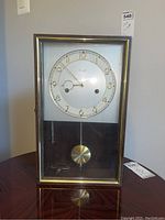 Front view of the rectangular pendulum wall clock showing the wooden frame, glass front, clock face with gold numerals, and brass pendulum with winding key inside.