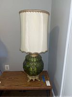 Full view of vintage lamp on wooden table with cream tall fabric shade and green glass base with gold-tone metal footed stand.