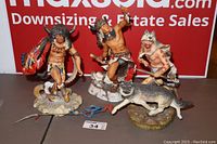 Overview of three Native American-themed statues: Spirit of the Sioux, Wolf Runner, and Buffalo Dancer placed on table against sale signage.