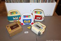 Five vintage Fisher Price cassette players for children displayed on table: cream/teal, white/red, white with colorful buttons, beige-brown, and white/grey with colored buttons.