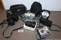 Lot of cameras, two camera bags, and a camera lens, displayed on table.