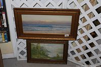Photo showing two vintage framed artworks stacked against a white lattice backdrop, the larger seascape on top and smaller landscape below. Both frames are brown wood with ornamental carving.