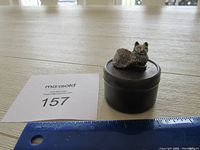 Small round metal tin with a detailed metal cat figurine sitting on the lid, placed beside a ruler and auction number card.