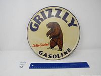 Front of the round metal sign showing the bear and the text 'GRIZZLY GASOLINE' and 'Dubbs Cracked'. A blue ruler beside the sign for scale.