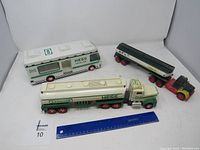 Three Hess toy trucks including white and green gasoline tanker semi-truck, black tanker trailer with red wheels, and white RV camper truck.