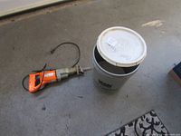 Photo of an electric jigsaw next to a gray bucket with white lid on a floor.
