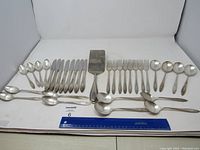 Full set of flatware arranged neatly including spoons, knives, forks, and serving spatula along with a ruler for size reference.