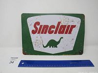 Full view of rectangular Sinclair metal sign with prominent text and dinosaur logo alongside a ruler for scale.