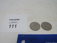Two 1776-1976 Bicentennial half dollar coins placed on a flat surface next to a ruler for scale and a MaxSold lot card.