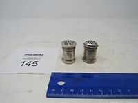 Two small cylindrical sterling silver salt and pepper shakers placed on a white surface with a blue ruler for size reference. Both show signs of tarnishing.