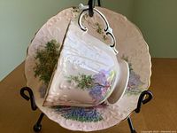 Teacup and saucer set displayed on a stand showing side profile of cup and front of saucer with country scene and floral painted details