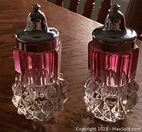Pair of cranberry glass salt and pepper shakers shown on wooden surface featuring tiara style silver tops and diamond point cut bases.