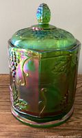 Front and side view of the green iridescent carnival glass canister showing the grape cluster and leaf pattern embossed on the body.