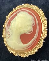 Front view of left facing cameo brooch showing white carved woman profile on reddish background with detailed gold metal frame
