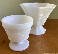 Two white milk glass pedestal dishes shape, grape vine pattern visible