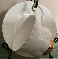 Side view of the white porcelain teacup with gold trim resting on the matching saucer, showing scalloped edges and gold accents on cup rim, handle, and base.