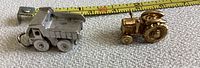 Side view of silver dump truck charm and gold tractor charm with measuring tape showing size