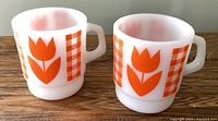 Two milk glass mugs side by side showing orange tulip and checkered design on white background.