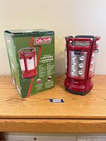 Photo of one red Coleman LED Quad Lantern next to its original green box with packaging graphics and text.