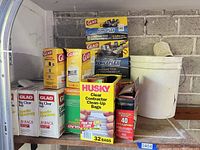 Clear shot of various boxes of Glad and Husky garbage bags of different colors and sizes, with white bucket on right side containing white towels.