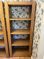 Two identical oak shelving units side by side against floral wallpaper