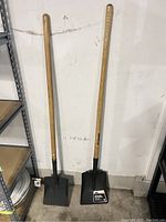 Photo of two shovels with wooden handles and square metal blades standing upright against a wall.