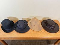Four specialty hats displayed side-by-side: black top hat, black safari hat, tan woven safari hat, and dark brown leather hat with braided band on wooden surface.