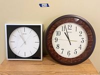 Two Seiko wall clocks side by side on a wooden surface: a round clock with brown marbled frame and a square clock with black and white frame.