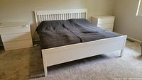 Front view of king size white wooden bed frame with bedding and two white dressers in a carpeted bedroom.
