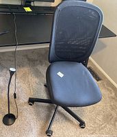 Black computer desk chair with mesh back and cushion seat next to black LED lamp with flexible neck on carpeted floor.