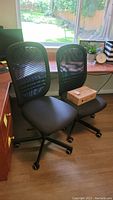 Two black computer chairs with mesh backs and padded seats placed side by side near a window and desk.
