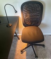 Black mesh back desk chair with padded seat next to black desk with lamp on top.