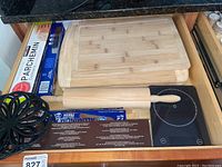 Drawer containing three wooden cutting boards, wooden rolling pin, digital kitchen scale, parchment paper, aluminum foil, and a black storage rack.