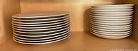 Photo of eleven 10-inch Grangehouse by Kennex USA plates stacked on the left and fifteen 8-inch IKEA plates stacked on the right in a wooden cabinet.