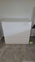 Full frontal view of the white three-drawer dresser standing on carpeted floor against a white wall.