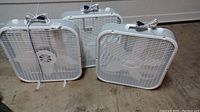 Three white Lasko electric box fans grouped on a sawdust-covered floor showing front view and cords on top.