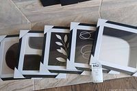 Five framed abstract art pieces stacked on a tiled floor showing various abstract shapes and designs in black frames with white mats.