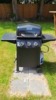 Front view of black Dyna-Glo propane BBQ grill on wheeled cart placed on patio stones with lawn background.