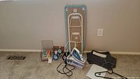 Wide shot showing the ironing board, iron with cord, hair dryer, toiletry bag, bath towels, and hangers on carpeted floor.