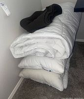 Stacked three white king-sized pillows and the comforter with a folded grey throw blanket on top, all folded and placed on carpet against white wall.