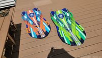 Two Sno-Storm snow sleds side by side on a wooden deck, one orange-blue and the other green-blue, showing overall look and size.